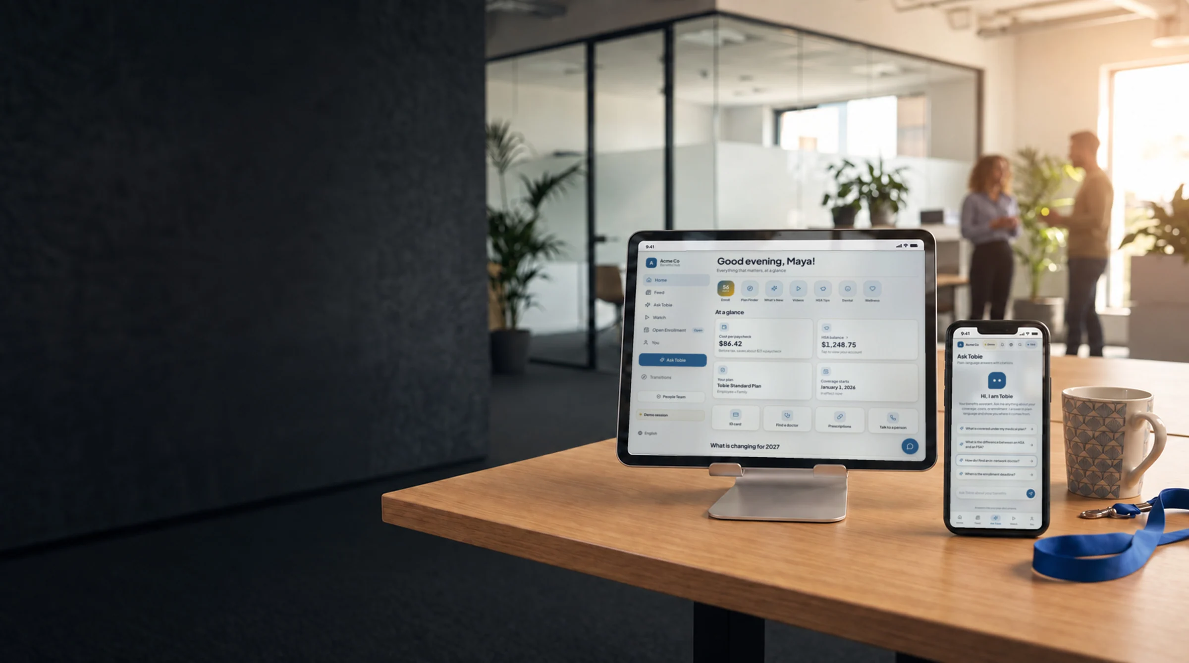 A tablet and a phone on a desk in an open-plan office: the benefits hub home on the tablet, Ask Tobie on the phone