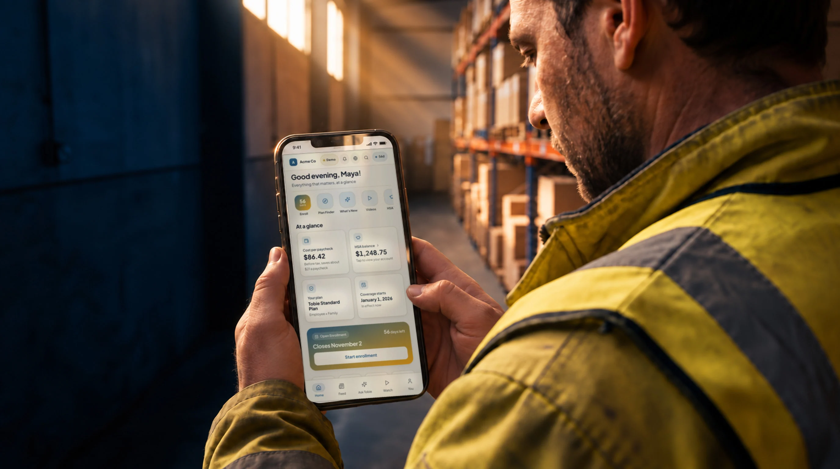 A warehouse associate on a break in a racking aisle, reading the benefits hub on his phone