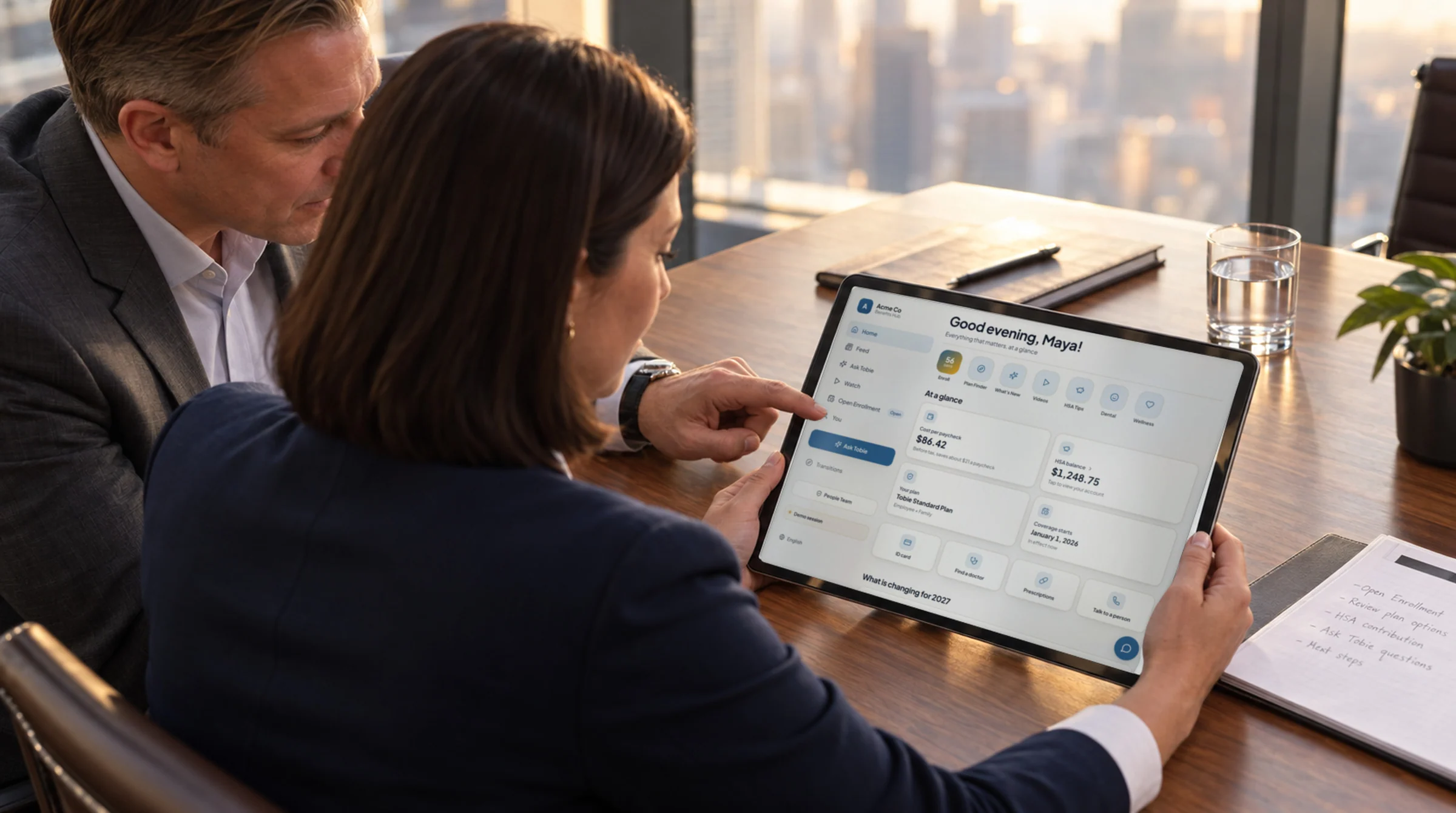 A benefits broker and an HR director reviewing the benefits hub on a tablet in a glass-walled meeting room