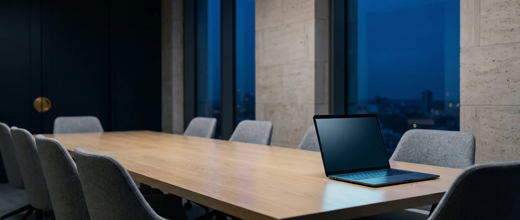 A laptop on a long boardroom table in a quiet office at dusk, city lights beyond the glass